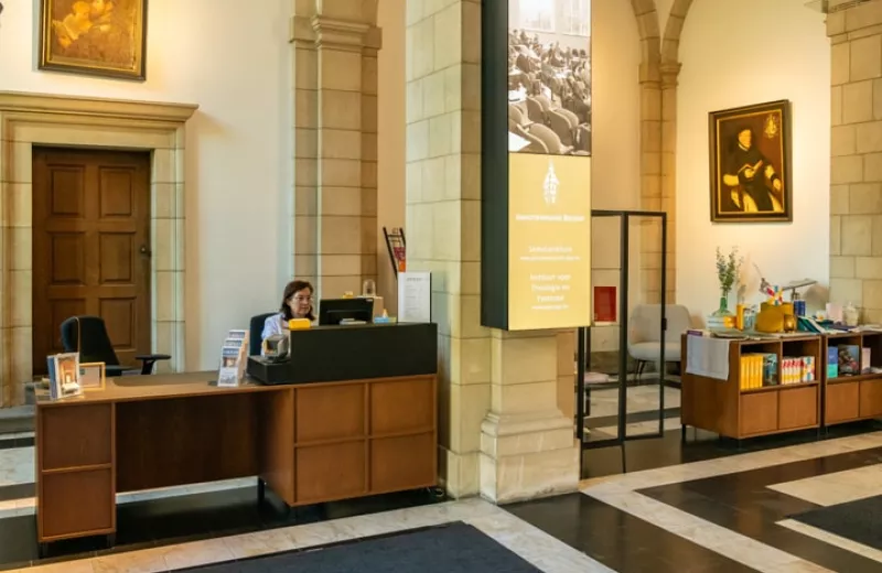 Mostrador de recepción y vestíbulo con columnas de piedra, muebles de madera, cuadros enmarcados en las paredes y un gran cartel vertical de exposición.