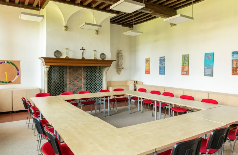 Sala de reuniones interior con una disposición en forma de U de mesas de madera clara y sillas rojas, una chimenea con una cruz y candelabros en la repisa, hornacinas arqueadas, vigas de madera a la vista en el techo y carteles verticales coloridos en las paredes blancas.