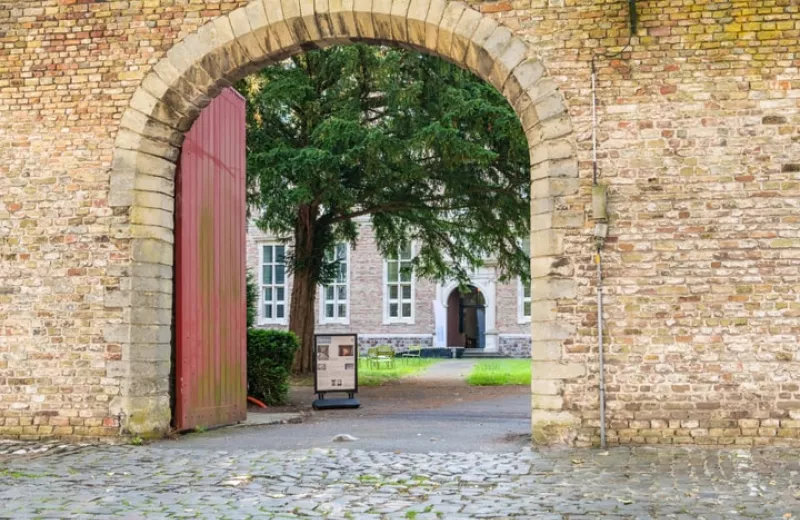 Arco de ladrillo con una gran puerta de madera roja que da a un patio empedrado con un árbol y un edificio blanco al fondo