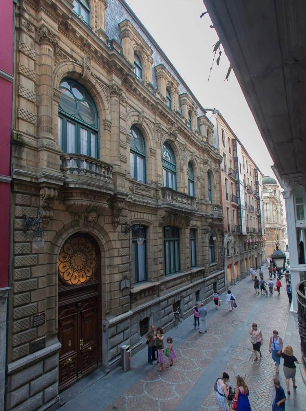 Rijkversierde historische stenen gevel met boogramen, decoratief metselwerk en een statige houten toegangsdeur met een ronde rozet, van bovenaf gezien langs een geplaveide voetgangersstraat met voorbijgangers.