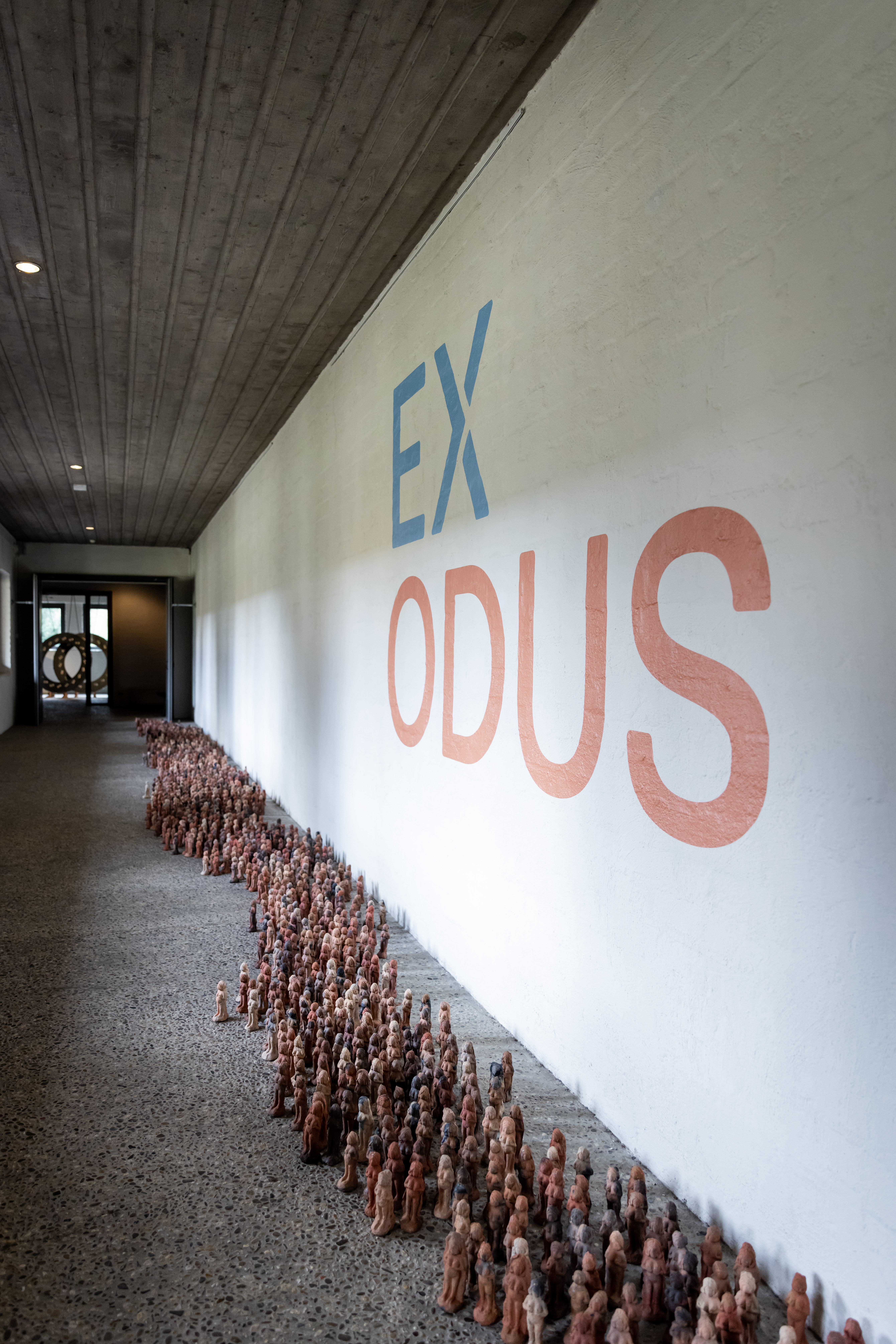 Een lange rij kleine beeldjes op de vloer naast een witte muur met het woord 'EXODUS' geschilderd in blauwe en oranje letters