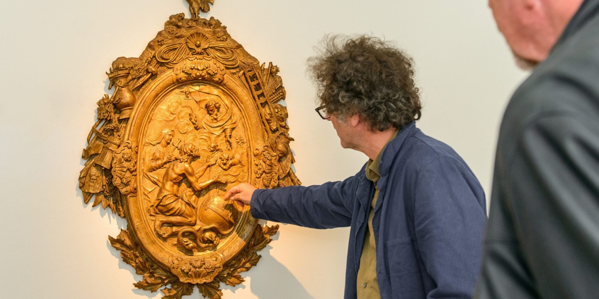 Persoon wijst naar een sierlijk gouden reliëf met Bijbelse taferelen op een witte muur in een museum