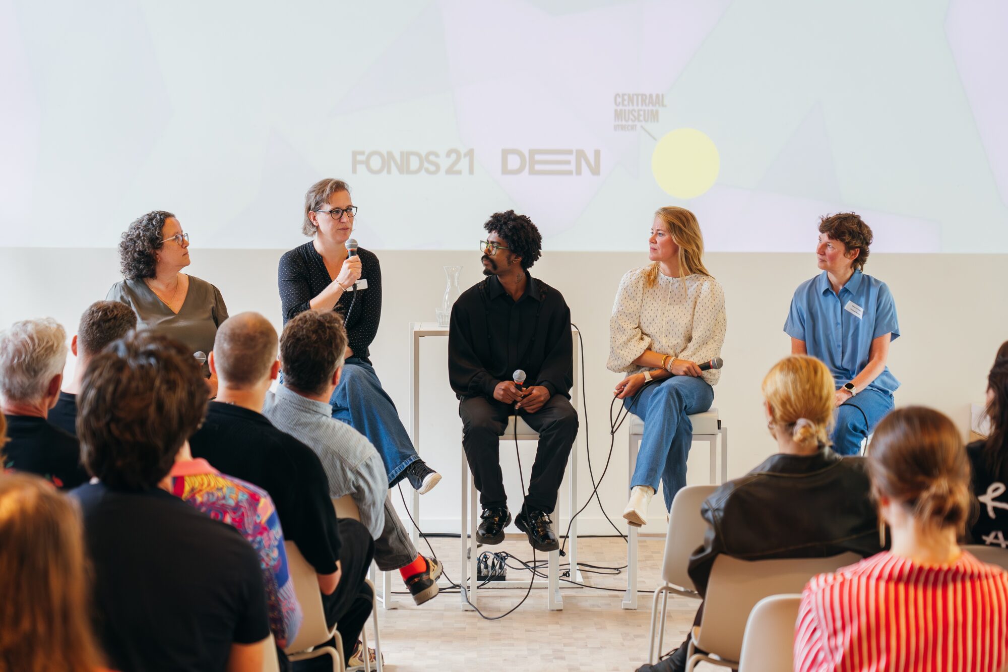 Paneldiscussie met vijf sprekers op het podium en publiek dat ervoor zit, achtergrond met FONDS21 en DIEN tekst