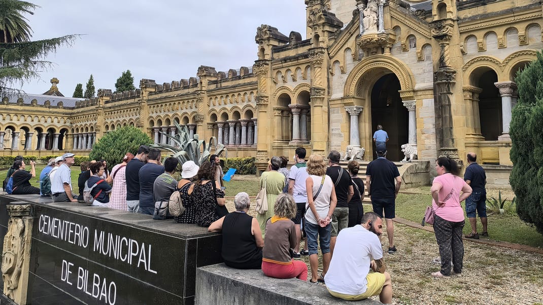 Tour group gathered in front of the ornate stone facade of Cementerio Municipal de Bilbao, with the cemetery name visible on a black granite sign in the foreground
