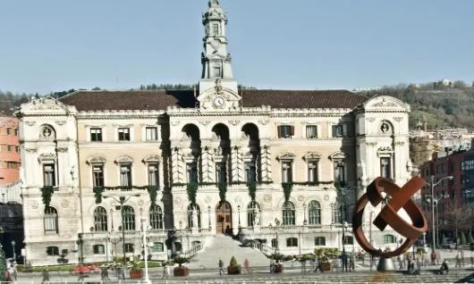 Ratusz w Bilbao, okazały neoklasycystyczny budynek z centralną wieżą zegarową i łukowatym portykiem wejściowym, z dużą rdzawą metalową rzeźbą na placu przed budynkiem