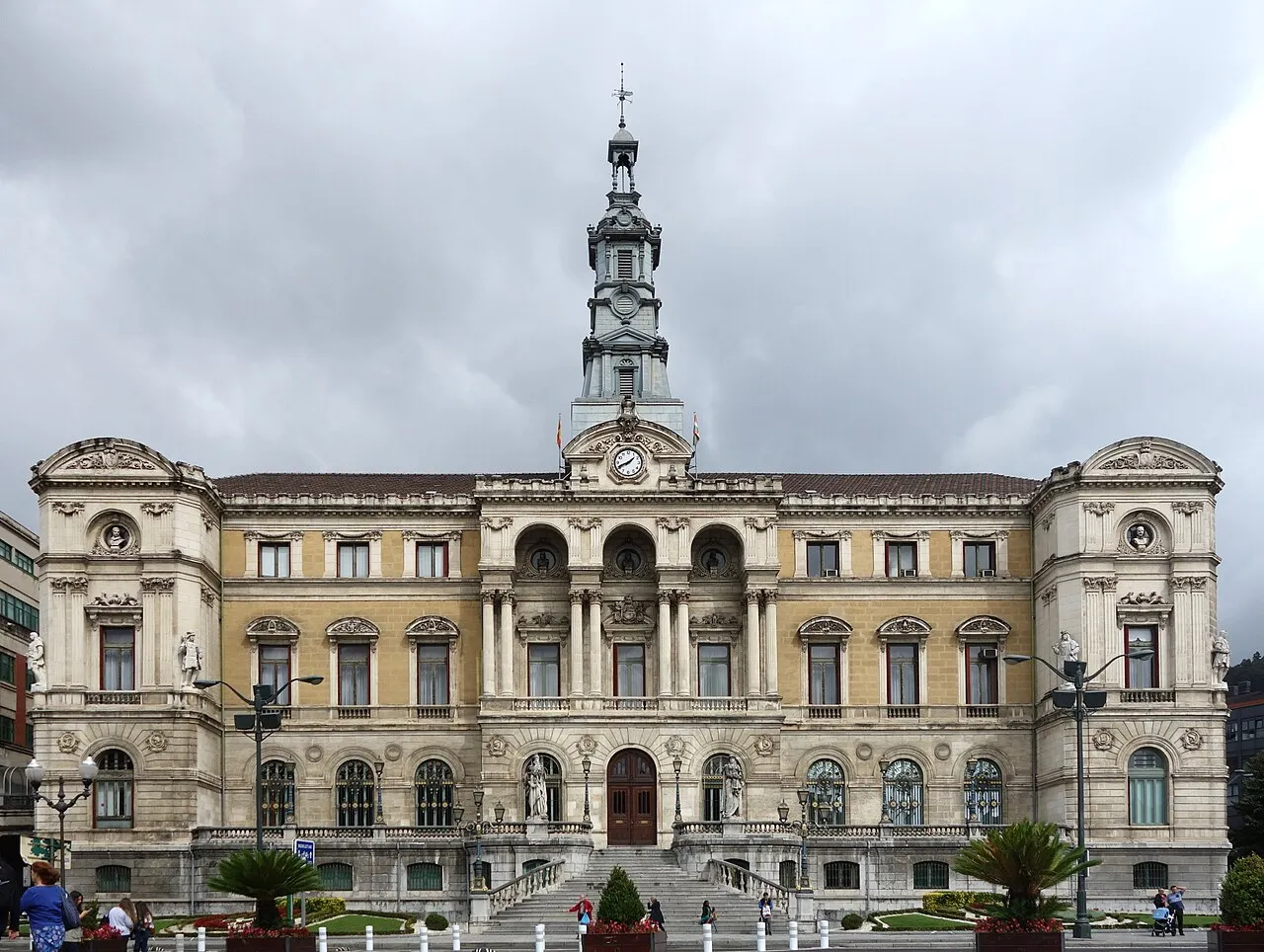 Wielki neoklasycystyczny budynek z ozdobną fasadą, centralną wieżą zegarową i łukowym wejściem, z ludźmi zgromadzonymi na placu przed nim