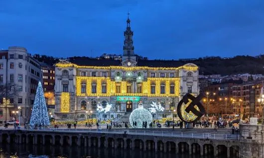 Ratusz w Bilbao oświetlony nocą złotym światłem, dekoracje świąteczne, w tym oświetlona choinka i figurki reniferów na placu, niebieskie niebo o zmierzchu