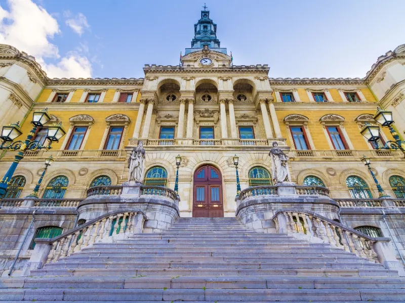 Frontowa fasada ratusza w Bilbao, ukazująca okazały żółty budynek w stylu neoklasycystycznym z centralną wieżą zegarową, łukowatymi oknami, posągami i szerokimi kamiennymi schodami prowadzącymi do głównego wejścia pod błękitnym niebem.