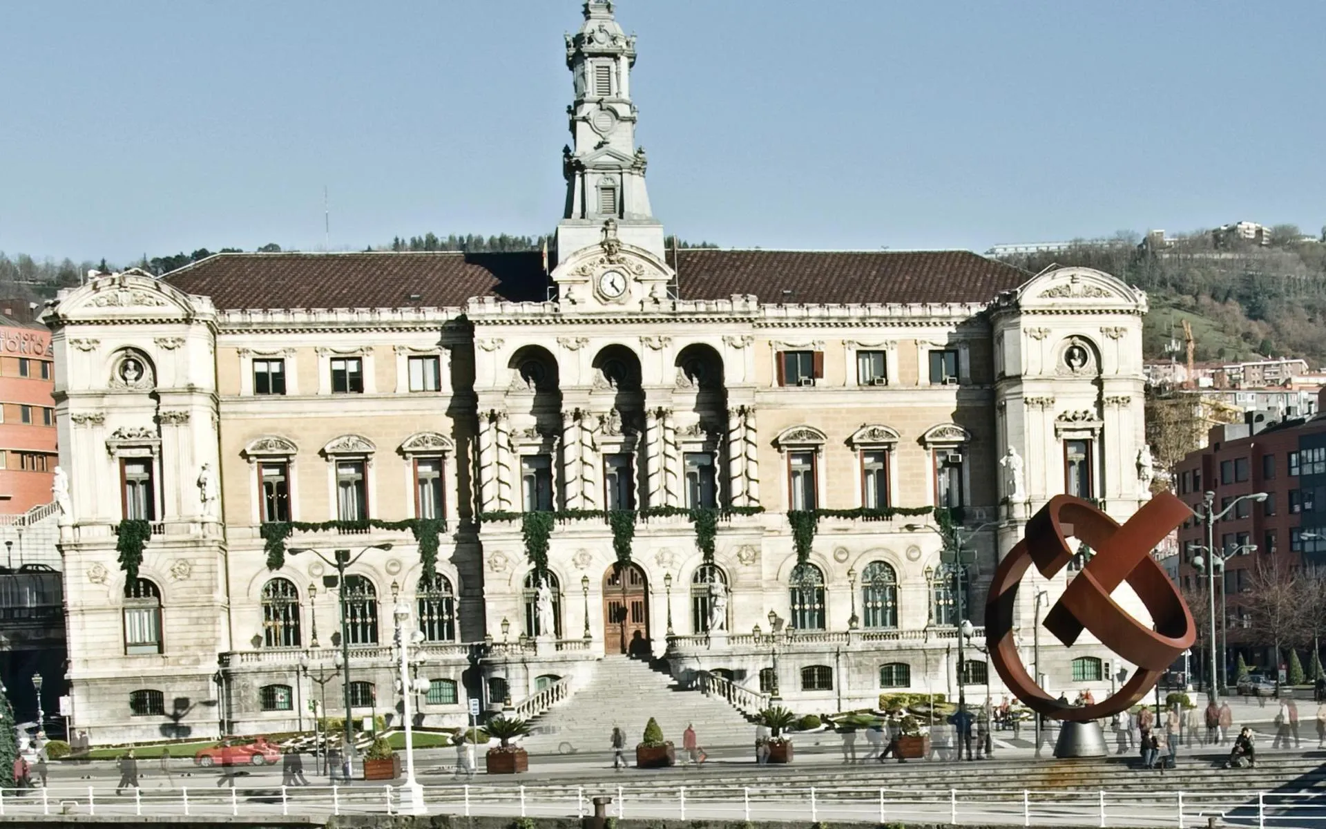 Szeroki widok z zewnątrz na ratusz w Bilbao, okazały, bogato zdobiony kamienny budynek z centralną wieżą zegarową i łukowym wejściem, z dużą rzeźbą z zardzewiałego metalu na pierwszym planie placu.