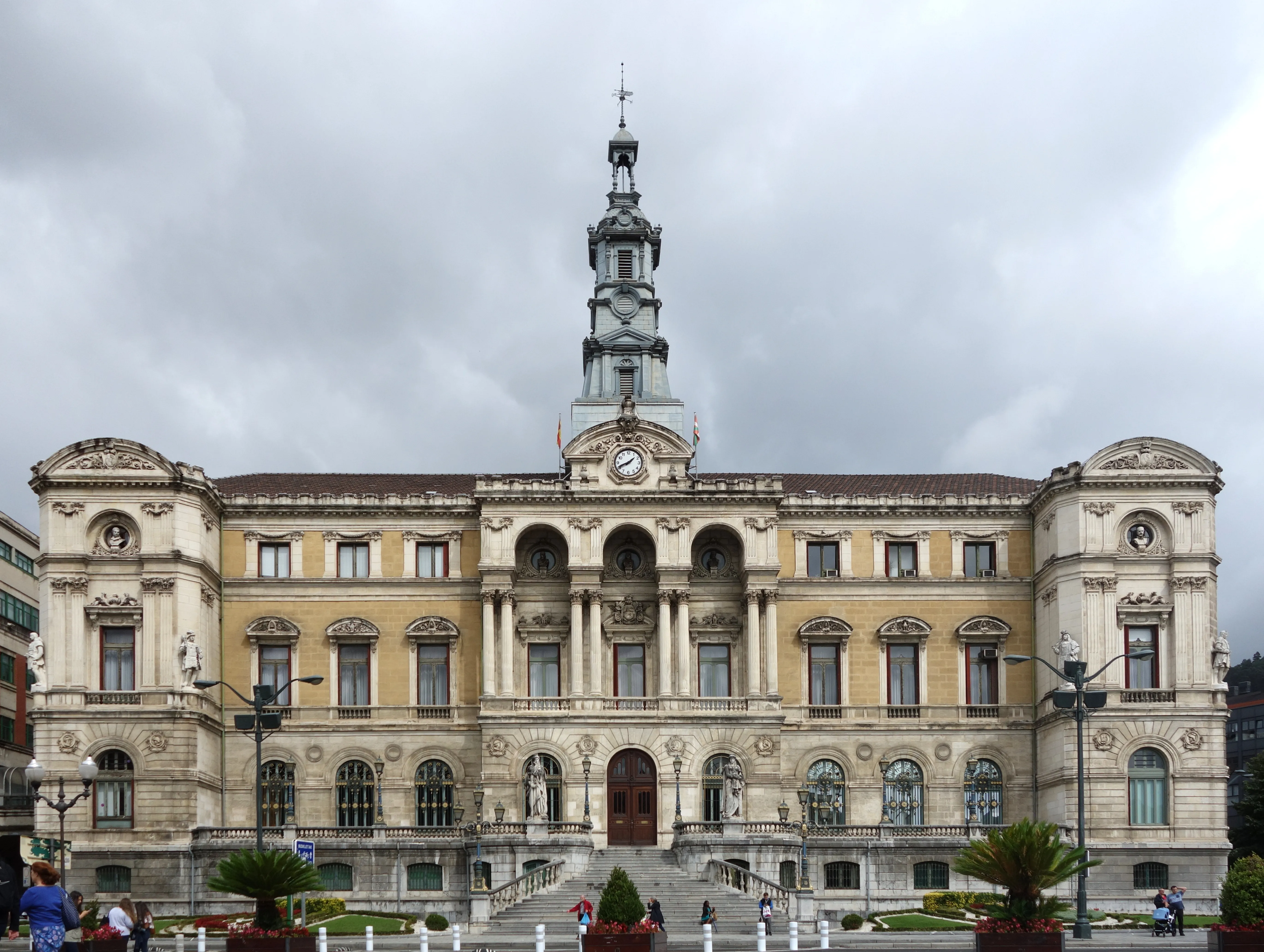 Fasada ratusza w Bilbao, przedstawiająca neoklasycystyczny budynek z centralną wieżą zegarową, łukowatymi oknami i zdobioną kamieniarką pod zachmurzonym niebem.