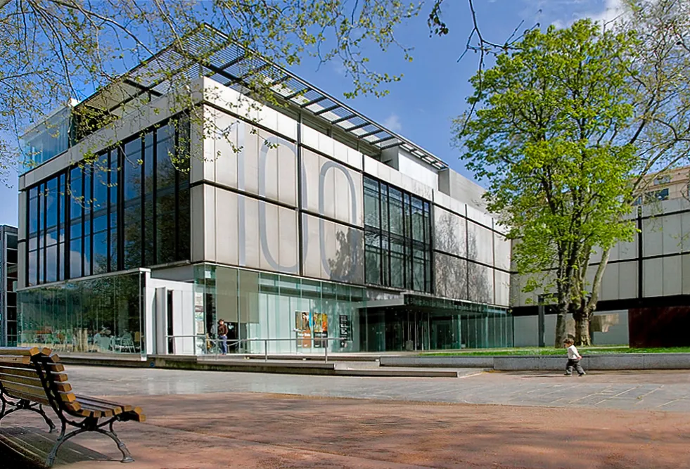 Moderne glazen en stalen gevel van het Museo de Bellas Artes de Bilbao met een houten bank op de voorgrond en groene bomen onder een blauwe lucht.
