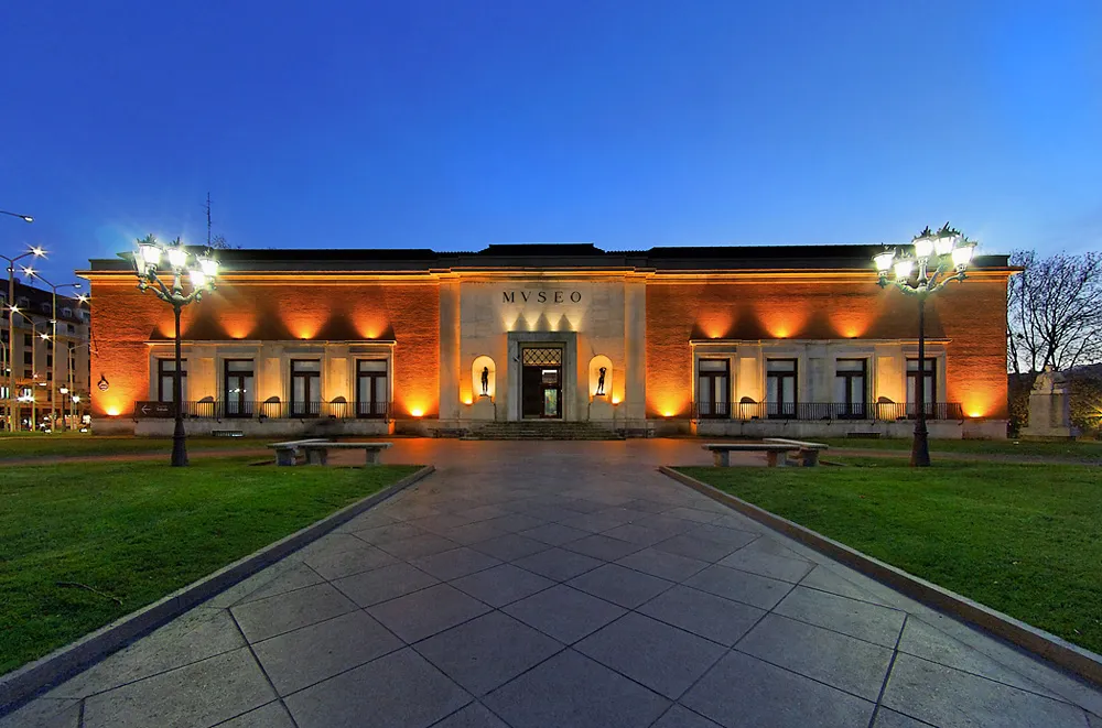 Verlichte gevel van het Bilbao Fine Arts Museum in de schemering, met warme oranje uplighting op een klassiek gebouw met het opschrift MUSEO, geflankeerd door sierlijke straatlantaarns en een geplaveid wandelpad.