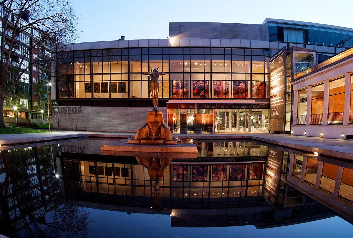 Exterieur van het Bilbao Fine Arts Museum bij schemering, met de verlichte glazen gevel, centrale fontein met standbeeld en een reflecterend bassin.