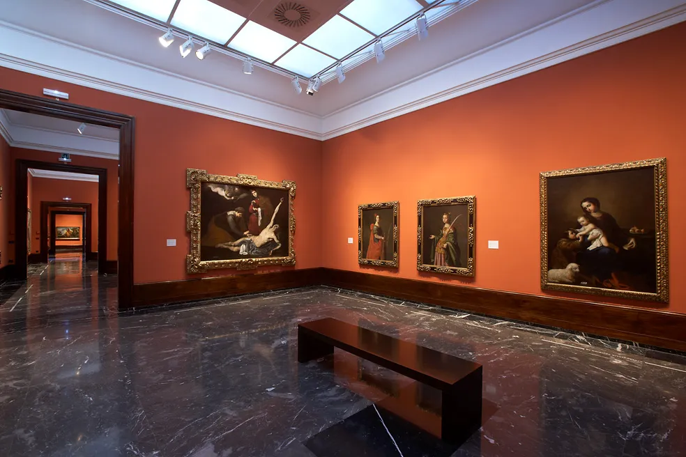 Interieur van een museumzaal met terracotta muren, donkere marmeren vloeren, een centrale houten bank en grote ingelijste schilderijen onder een lichtkoepel.