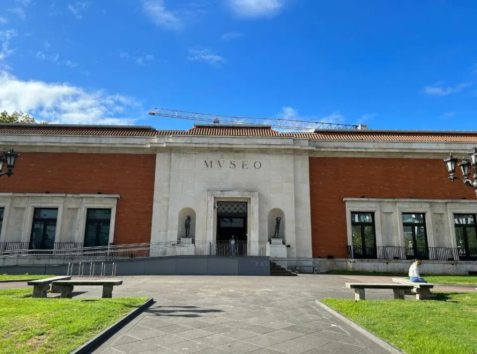 Rode bakstenen museumgebouw met een witte centrale portiek bij de ingang waarop MUSEO staat, stenen bankjes en een groen gazon ervoor onder een blauwe lucht