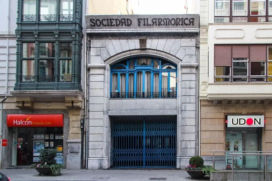 Fachada de piedra del edificio de la Sociedad Filarmónica que presenta una gran entrada arqueada con puertas de metal azul y marcos de ventana azules encima, flanqueada por escaparates vecinos.