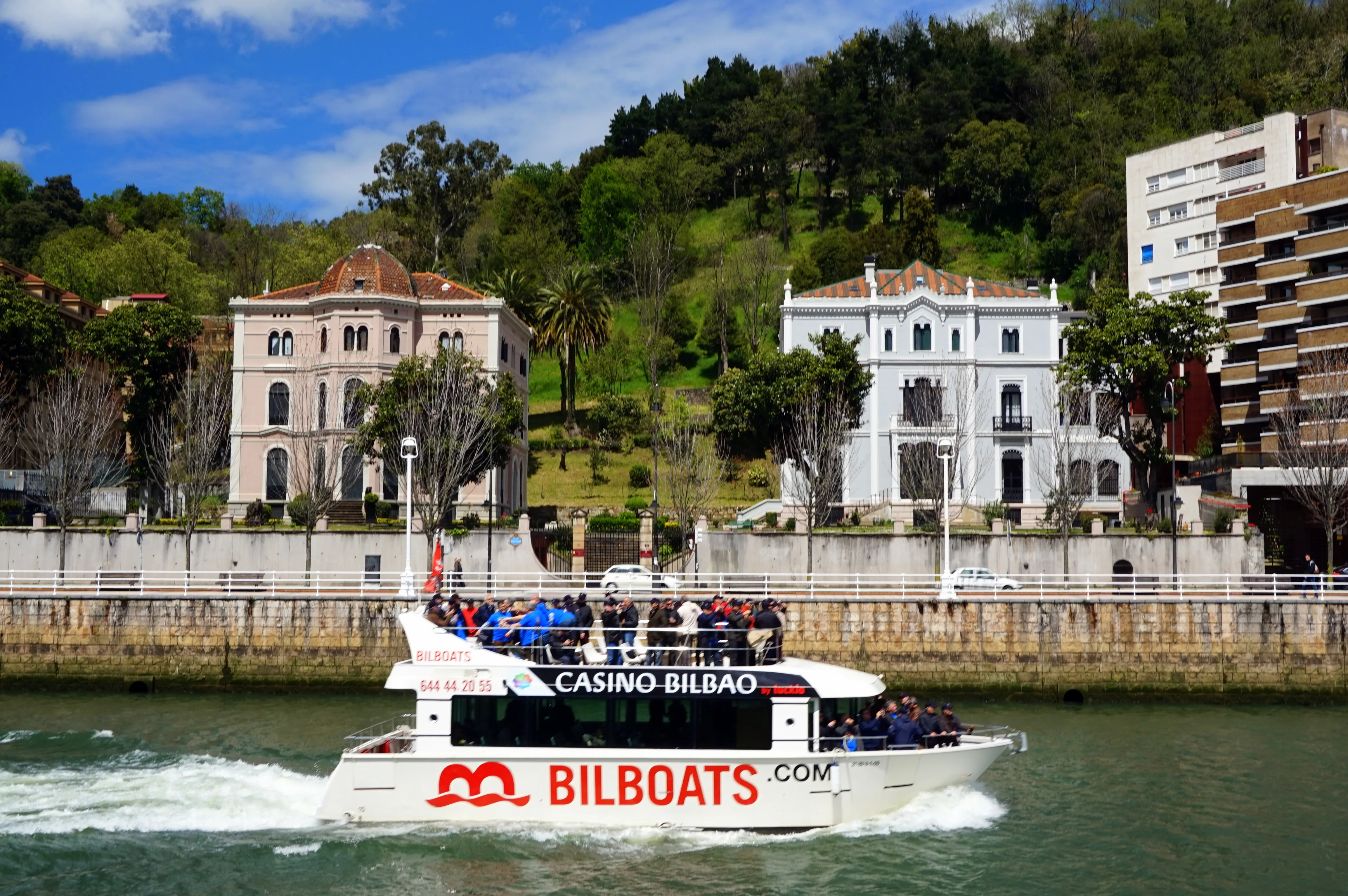 Biały statek wycieczkowy Bilboats.com z pasażerami na górnym pokładzie płynący rzeką Nervión w Bilbao, z zabytkowymi budynkami i zielonymi wzgórzami w tle.