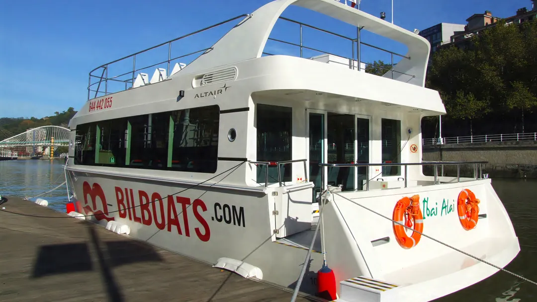 Biały katamaran z dużym czerwonym napisem 'BILBOATS.COM' na kadłubie, zacumowany przy nadbrzeżu, z pomarańczowymi kołami ratunkowymi na rufie i widocznym w tle mostem.