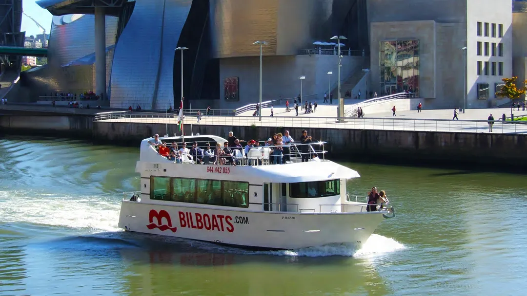 Biała łódź wycieczkowa firmy Bilboats.com z pasażerami na górnym pokładzie płynąca po ujściu rzeki w Bilbao, z widocznym w tle Muzeum Guggenheima w Bilbao.