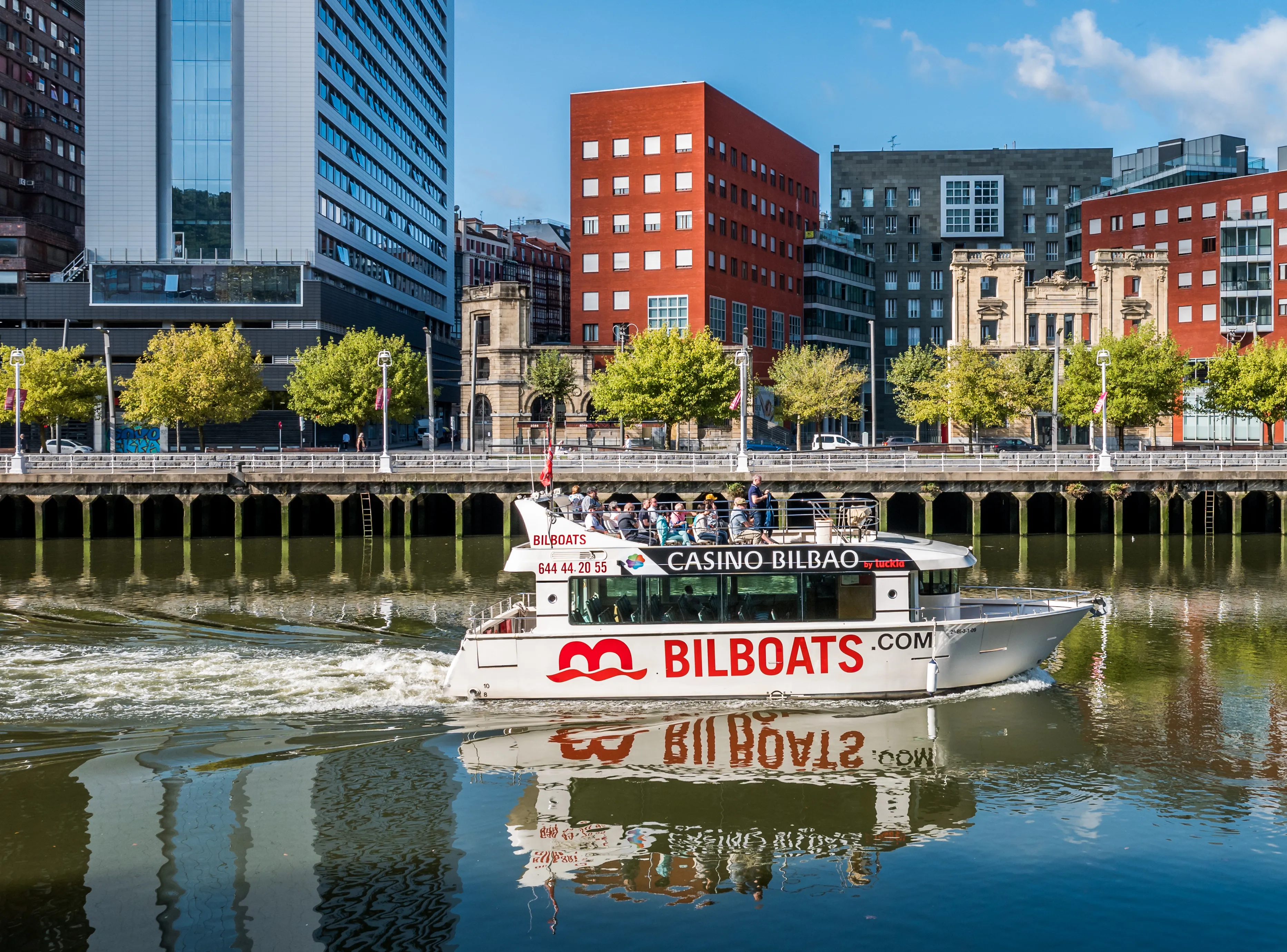 Biały statek wycieczkowy Bilboats z pasażerami płynie wzdłuż estuarium Bilbao, na tle nowoczesnych szklanych wieżowców i budynków z czerwonej cegły stojących wzdłuż brzegu rzeki.