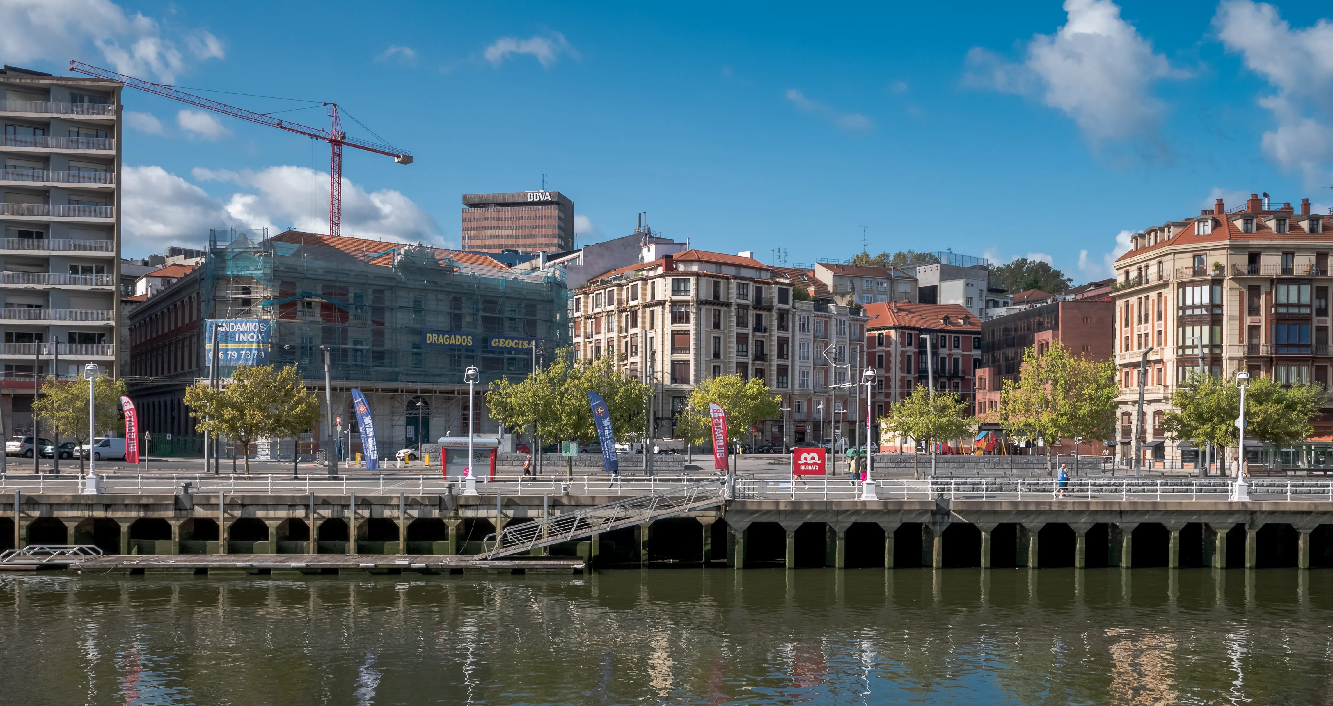 Szeroki panoramiczny widok na nabrzeże rzeki Bilbao Estuary, ukazujący wysadzaną drzewami promenadę, mieszankę zabytkowych i nowoczesnych budynków, dźwig budowlany oraz odbicia w wodzie pod błękitnym niebem.