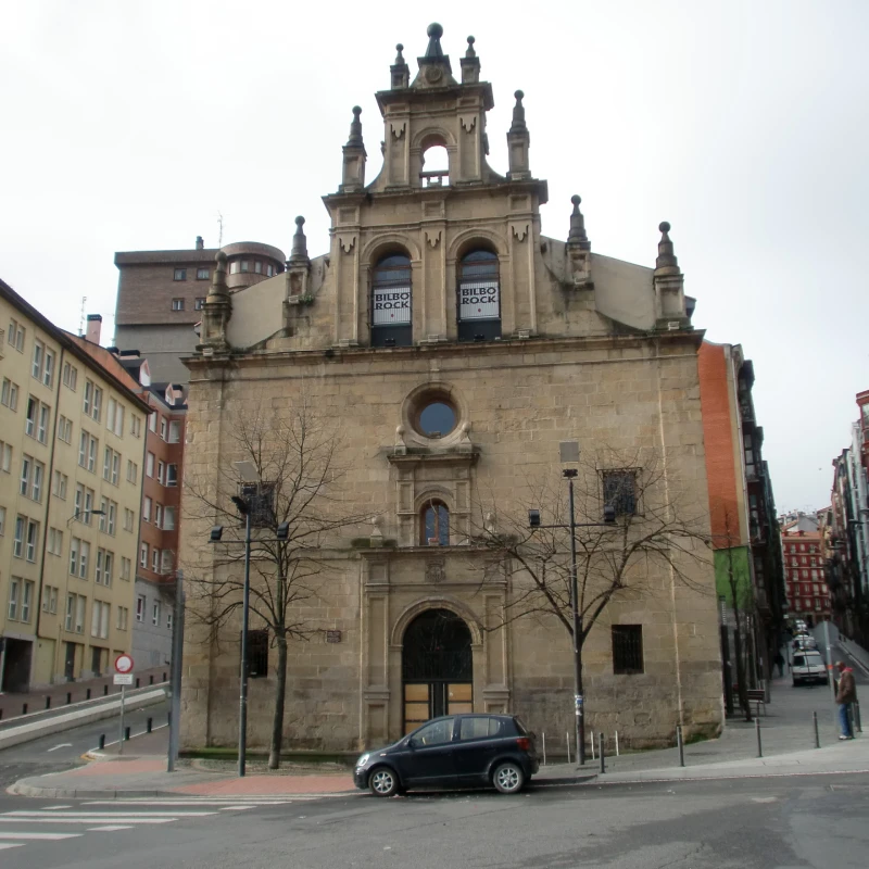 Fachada de iglesia barroca de piedra con torres gemelas, ventanas arqueadas con estandartes, un óculo circular y un pequeño coche negro aparcado delante en una calle de la ciudad.