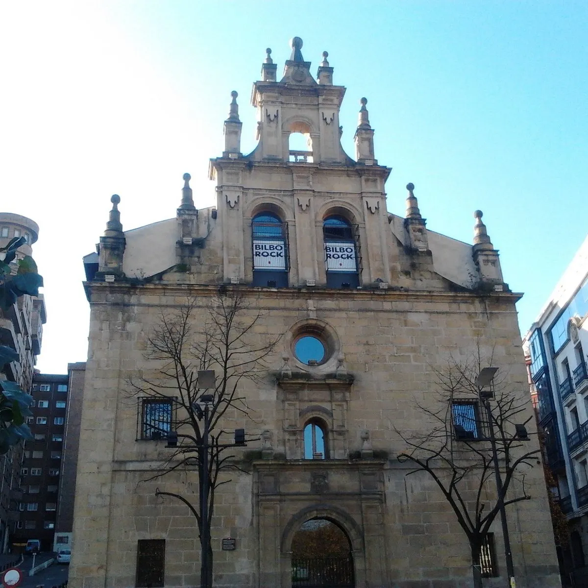 Fachada de piedra de un histórico edificio eclesiástico barroco con pancartas de Bilbo Rock en los ventanales arqueados superiores, flanqueado por árboles desnudos contra un cielo azul