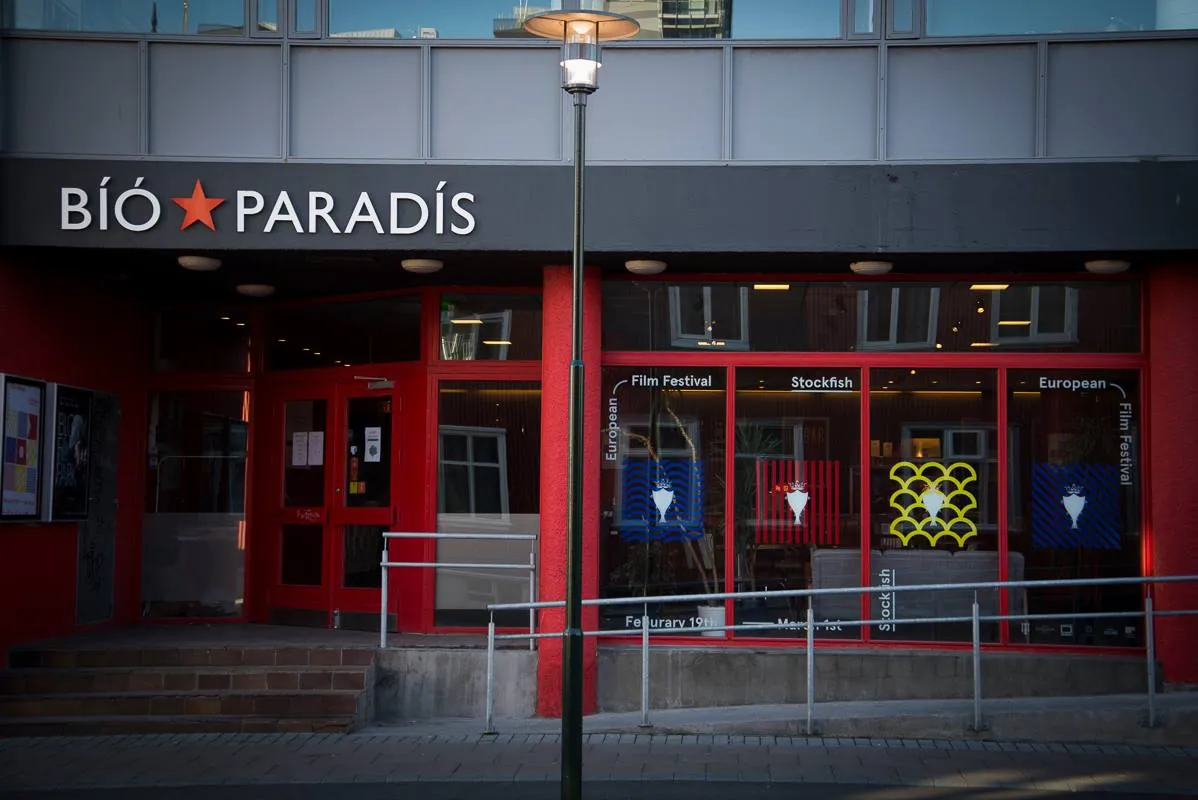 Fasada zewnętrzna kina Bíó Paradís z czarnym szyldem z białym napisem i czerwoną gwiazdą, czerwonymi ramami okien z plakatami filmowymi oraz latarnią uliczną przed budynkiem.
