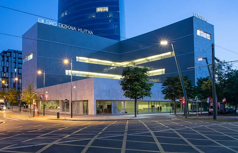 Buitenaanzicht van het Bizkaia Aretoa-gebouw in de schemering, met de hoekige betonnen gevel, verlichte horizontale ramen en het uithangbord van El Bao Bizkaia Kutxa boven een geplaveid plein.