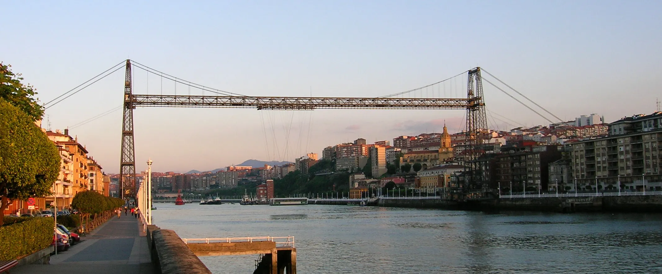 Amplia vista panorámica del Puente de Bizkaia, un puente transbordador que cruza la ría con torres de hierro y vía de góndola suspendida, flanqueado por edificios urbanos a ambas orillas al atardecer.