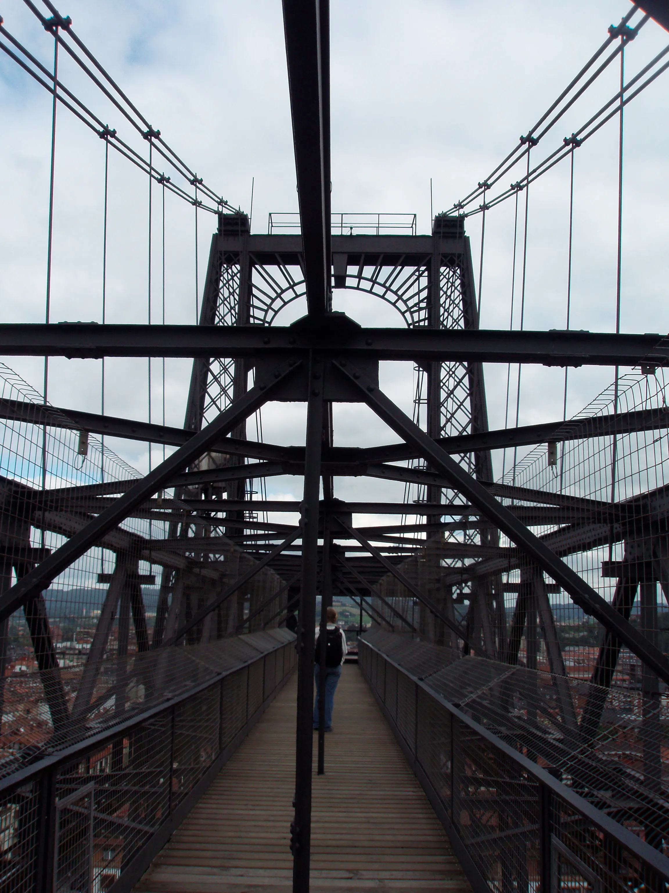Pasarela peatonal superior del Puente de Bizkaia mostrando torres de celosía de hierro, cables de suspensión y vigas transversales de acero con una persona caminando por la pasarela de madera