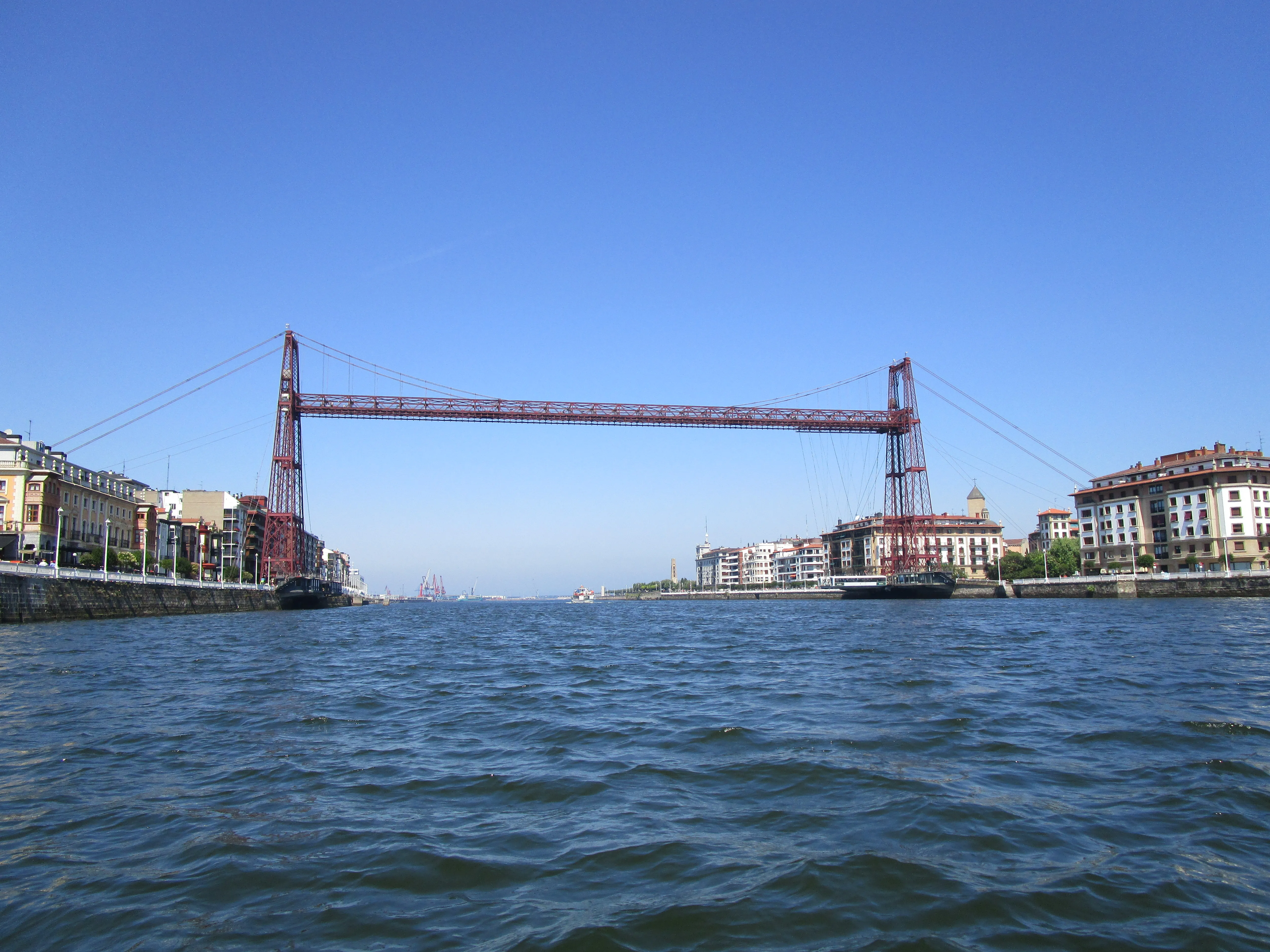 El Puente Bizkaia cruzando la ría entre Portugalete y Getxo, visto desde el agua bajo un cielo azul despejado con sus torres de hierro rojo y su estructura de góndola suspendida.