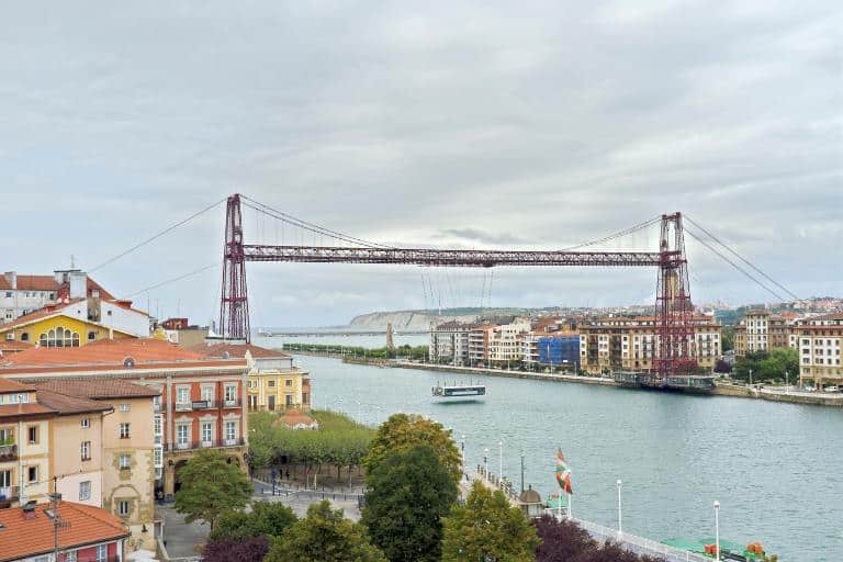 El puente transbordador de Bizkaia que cruza un estuario con torres de hierro rojo y una góndola suspendida, flanqueado por edificios coloridos en ambas orillas