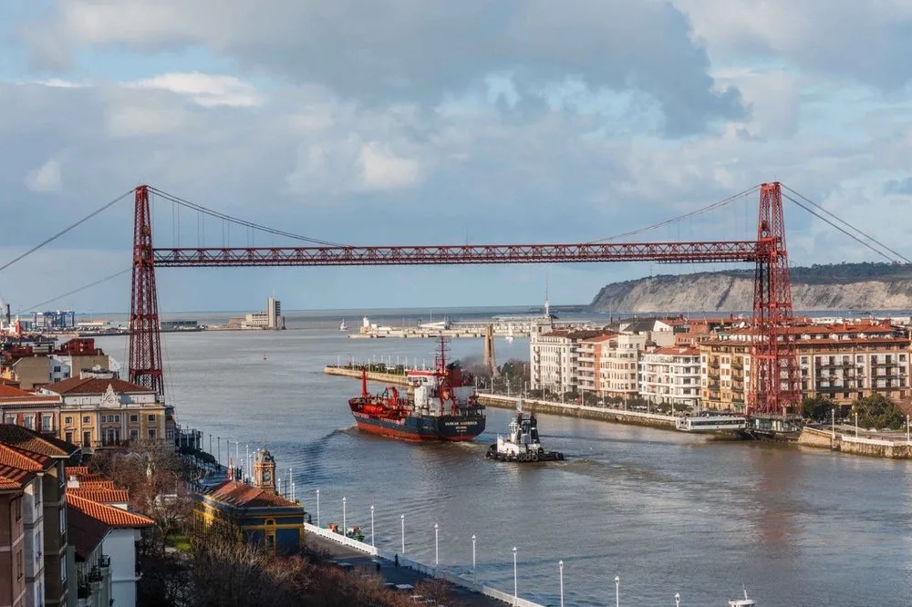 El puente transbordador de hierro rojo de Bizkaia sobre la ría del Nervión, con un buque de carga pasando por debajo y edificios a ambos lados de la orilla bajo un cielo nublado