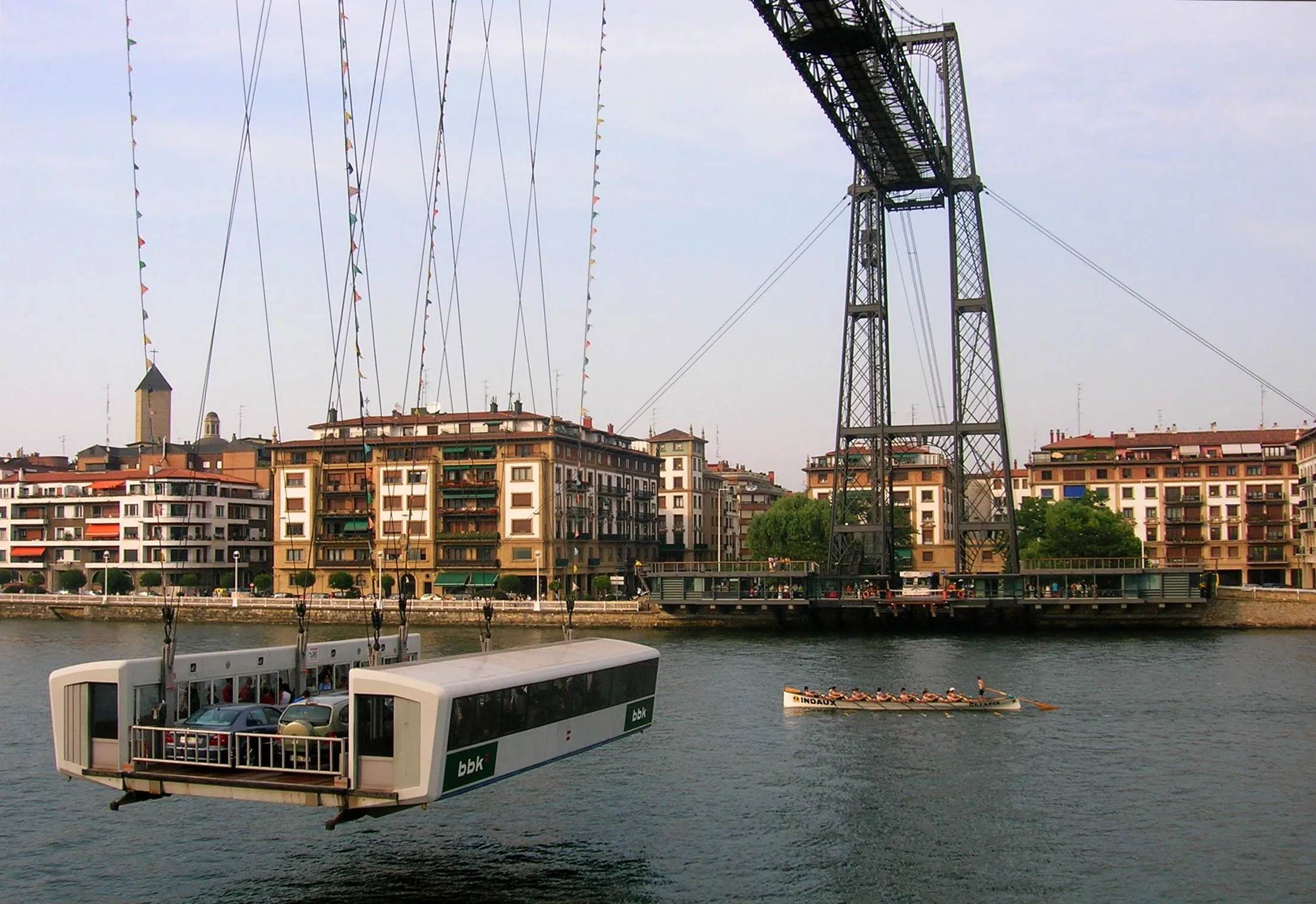 Góndola colgante del Puente Bizkaia cruzando la ría con su torre de hierro y los edificios de la orilla al fondo, y un bote de remos sobre el agua