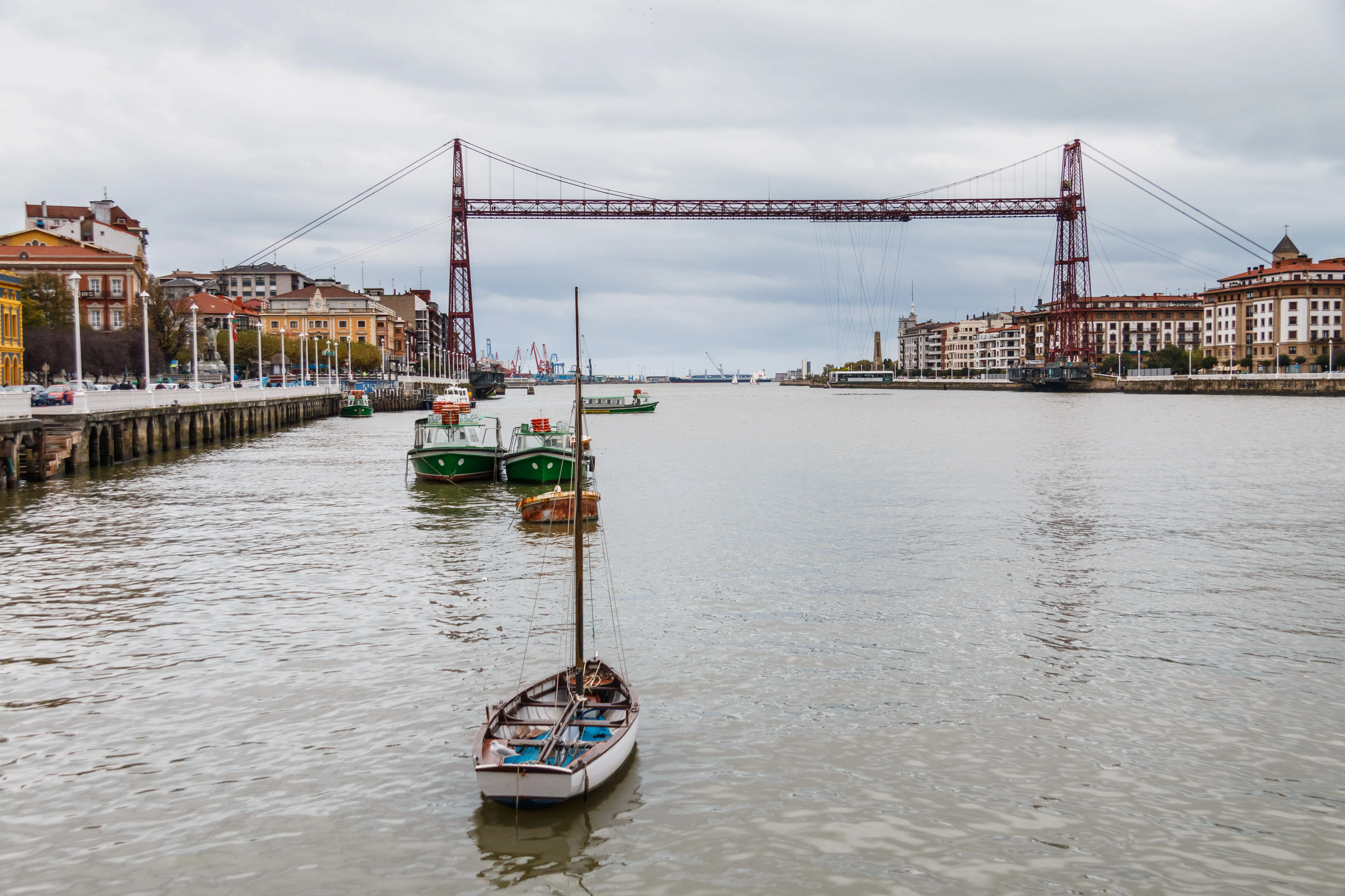 El puente transbordador de Bizkaia cruza la ría con sus torres de hierro rojo y su barquilla suspendida, con un velero en primer plano y edificios a orillas del agua a ambos lados bajo un cielo nublado.