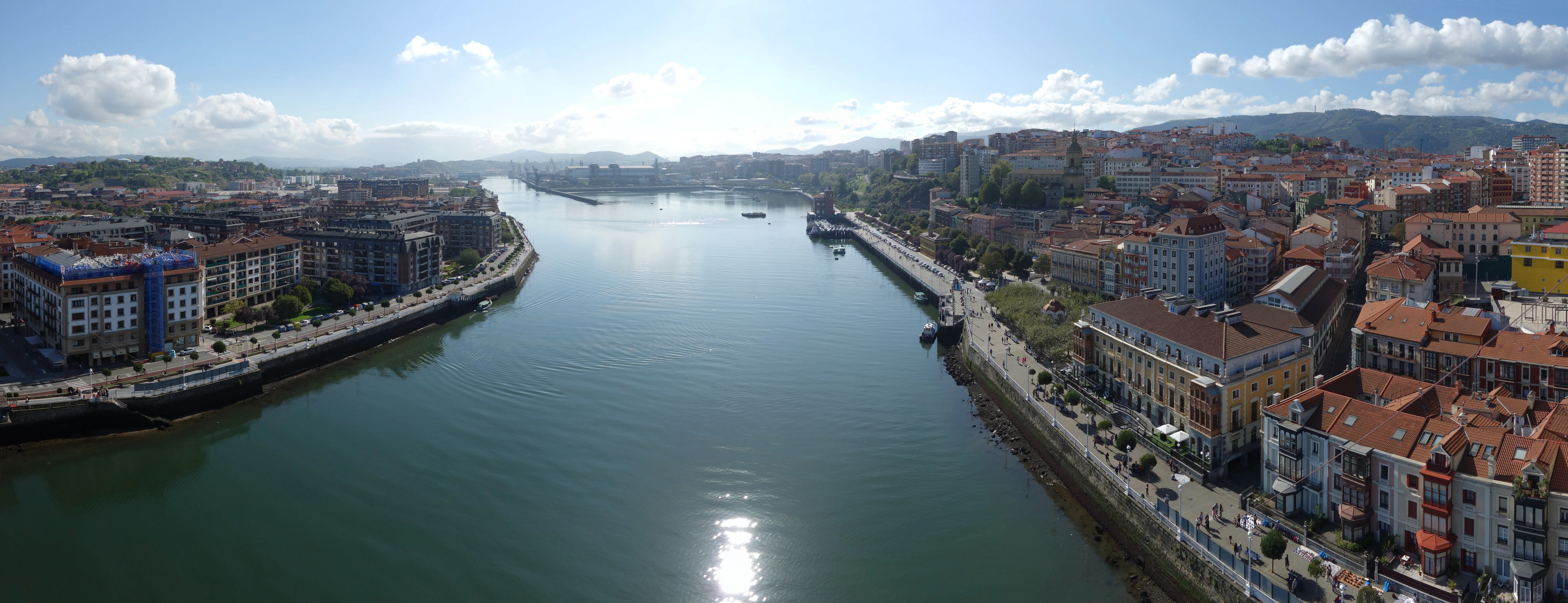 Vista panorámica del Puente de Vizcaya que cruza la ría del Nervión entre Portugalete y Getxo, con edificios urbanos a ambas orillas del río bajo un cielo despejado y el sol reflejándose en el agua.