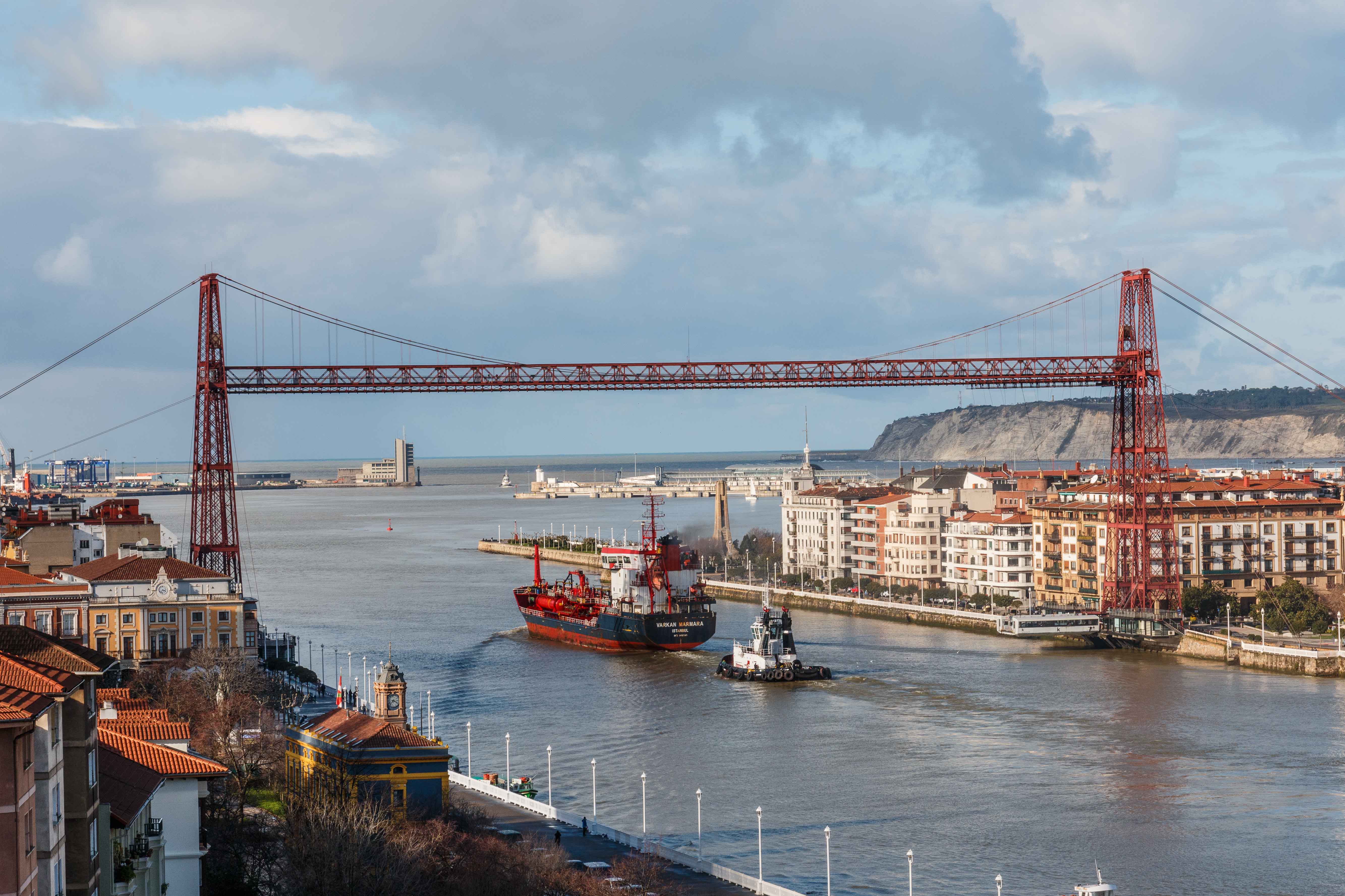 Puente transbordador de hierro rojo que cruza un amplio estuario con barcos pasando por debajo, flanqueado por pueblos costeros y acantilados bajo un cielo nublado.