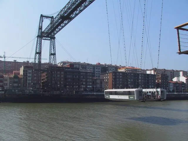 El puente transbordador de Bizkaia con su barquilla suspendida transportando un autobús a través de la ría, torres de celosía de hierro y edificios urbanos visibles al fondo bajo un cielo azul despejado