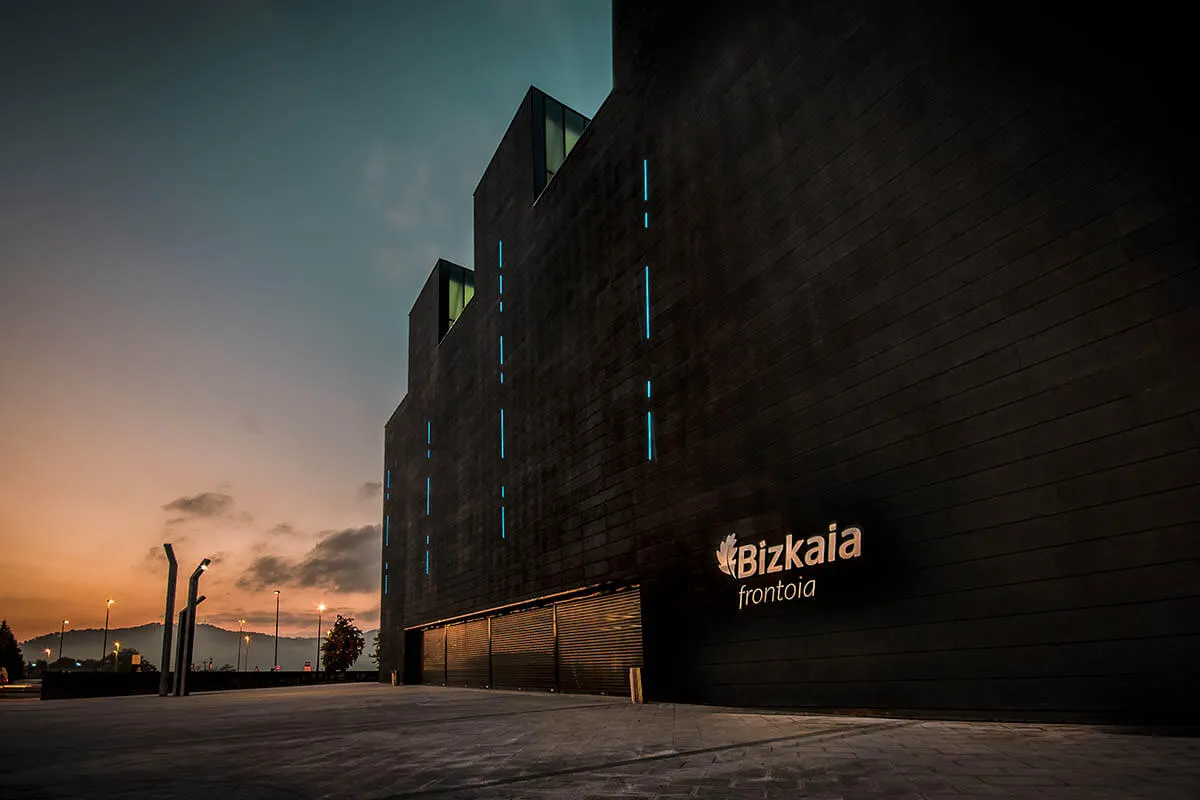 Dark modern building facade at dusk with illuminated 'Bizkaia frontoia' sign and vertical light strips against a sunset sky