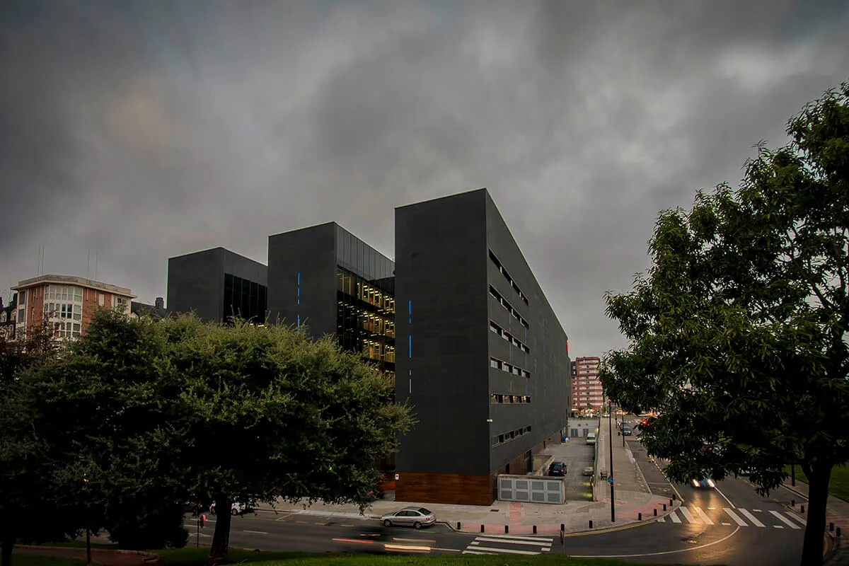 Modern dark gray multi-block building complex with large glass windows, framed by mature trees and a street with crosswalk under a dramatic cloudy sky