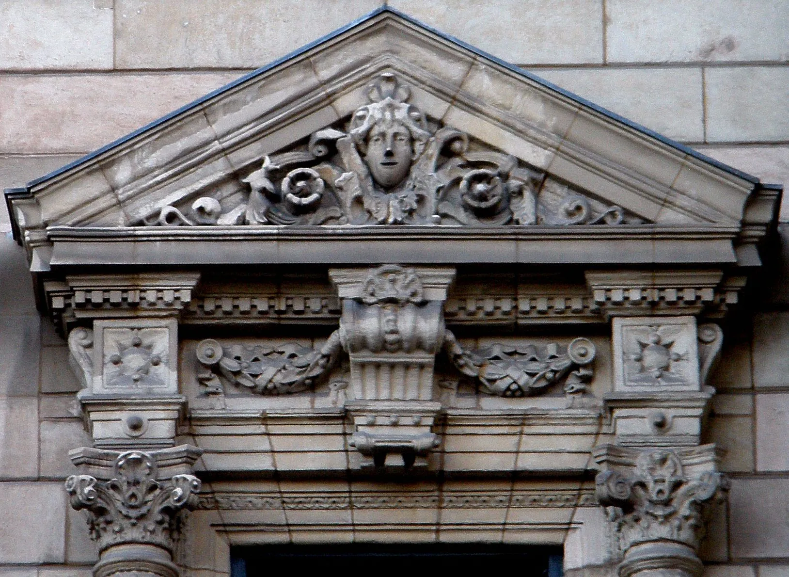 Ornate stone pediment with a carved mascaron face in the tympanum, decorative scrollwork, a central keystone, and Corinthian column capitals on the building facade