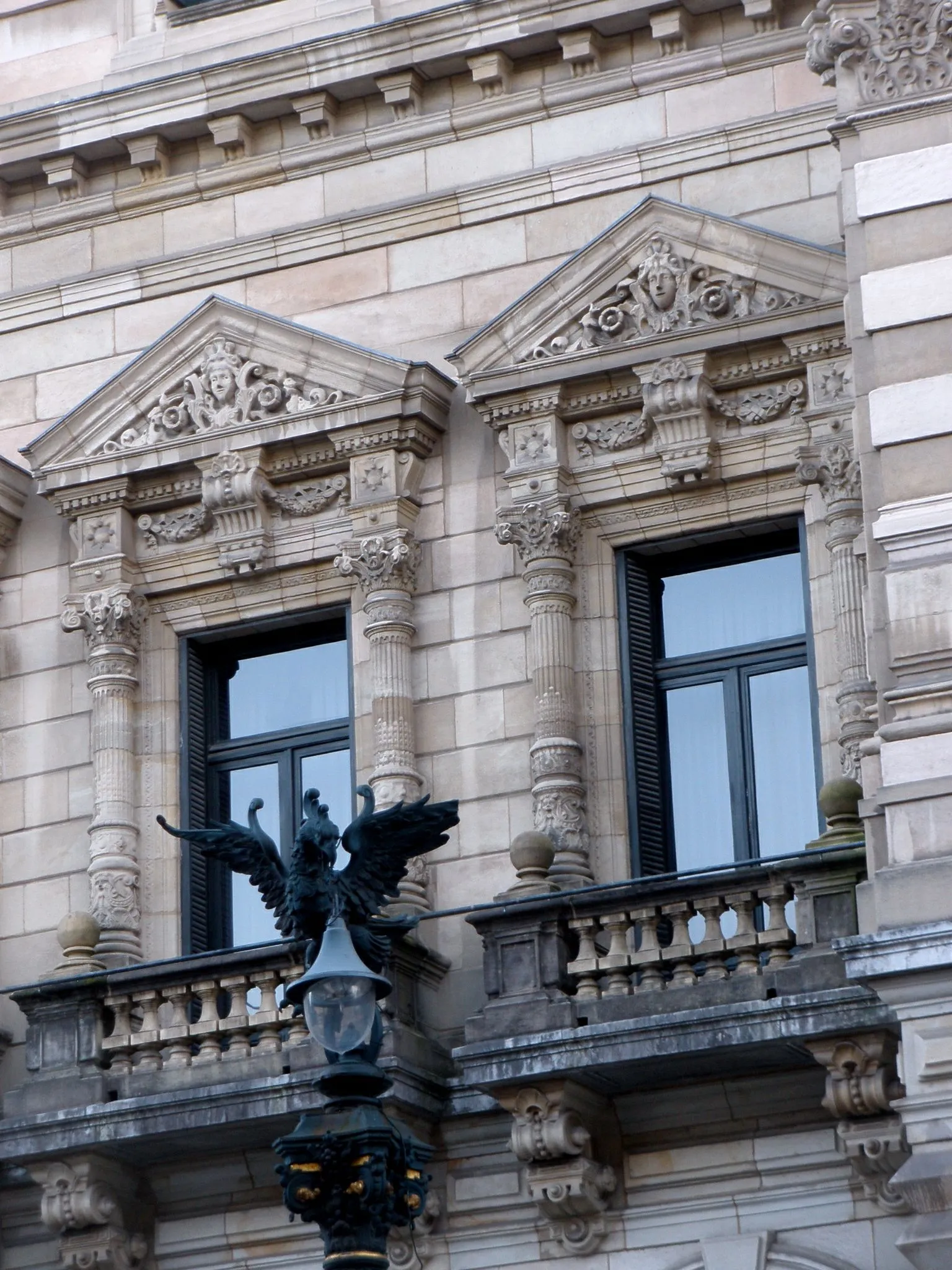 Ornate stone facade of the Bizkaia Provincial Council Palace showing two windows with sculpted triangular pediments, decorative columns, a balustrade, and a wrought-iron lamp post with a winged figure in the foreground