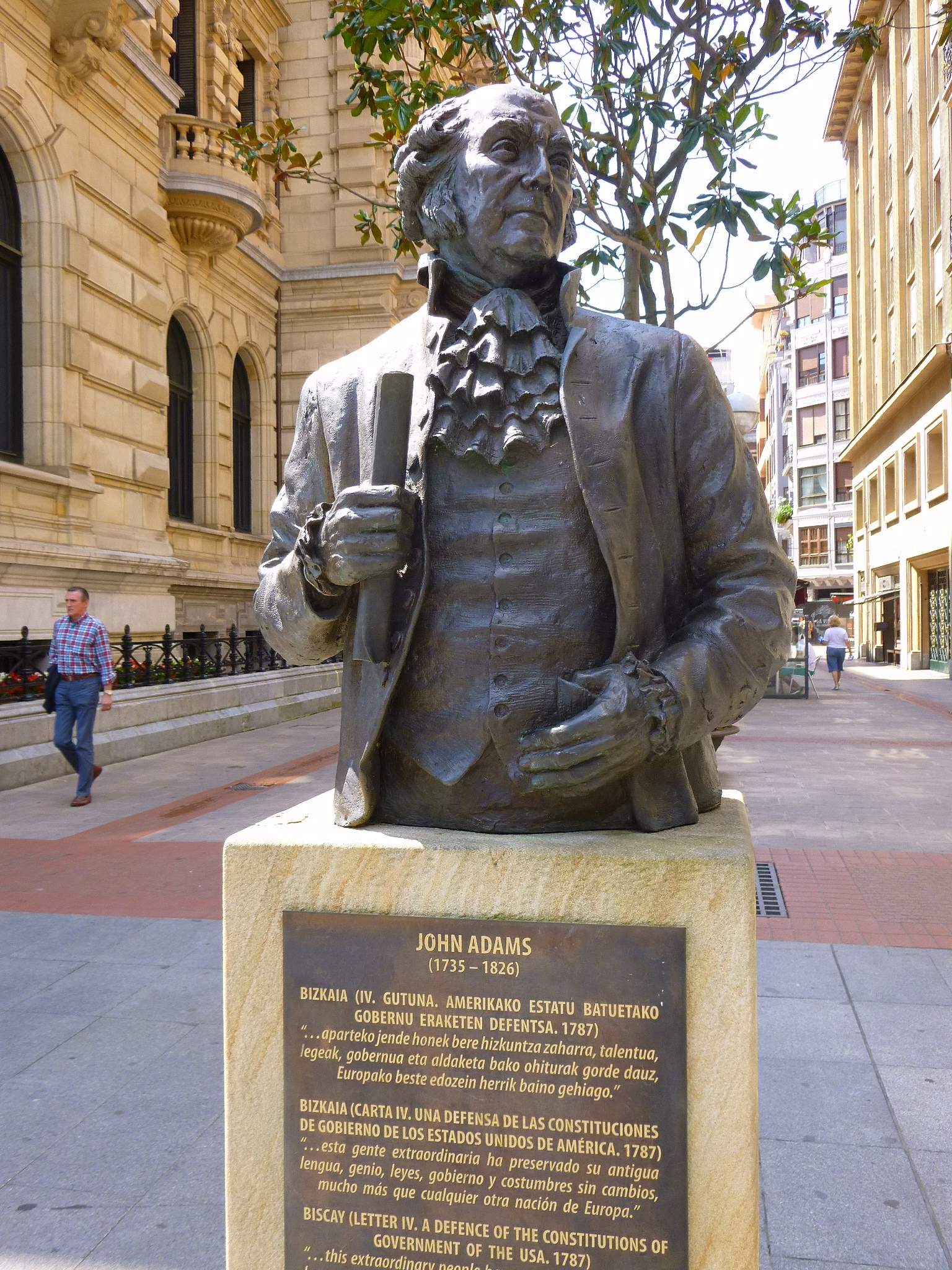 Bronze bust of John Adams on a stone pedestal with a multilingual commemorative plaque, set on a pedestrian street with an ornate stone building visible in the background.