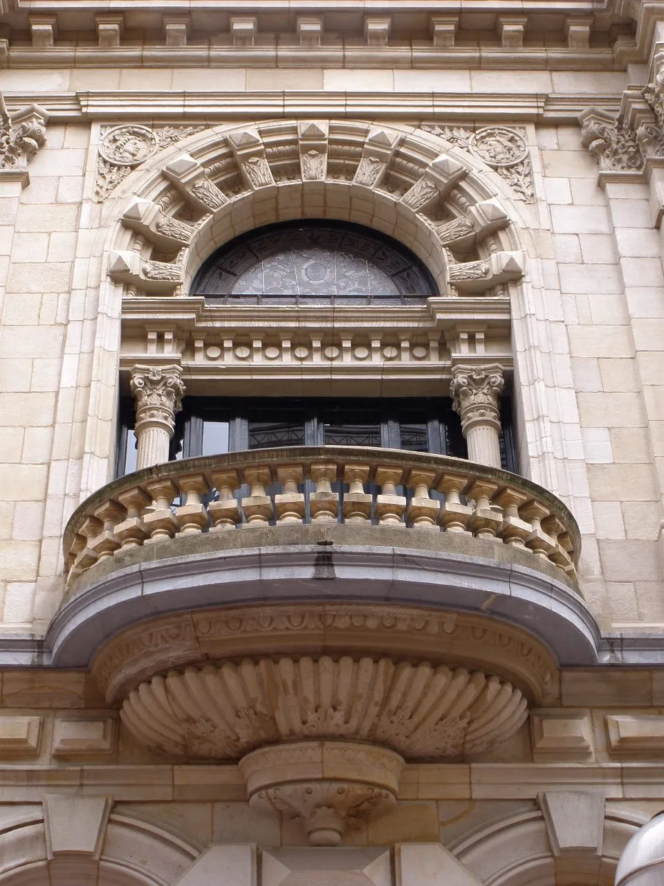 Ornate stone balcony with curved balustrade, Corinthian columns, decorative corbel, and a semi-circular arched window above on a historic building facade