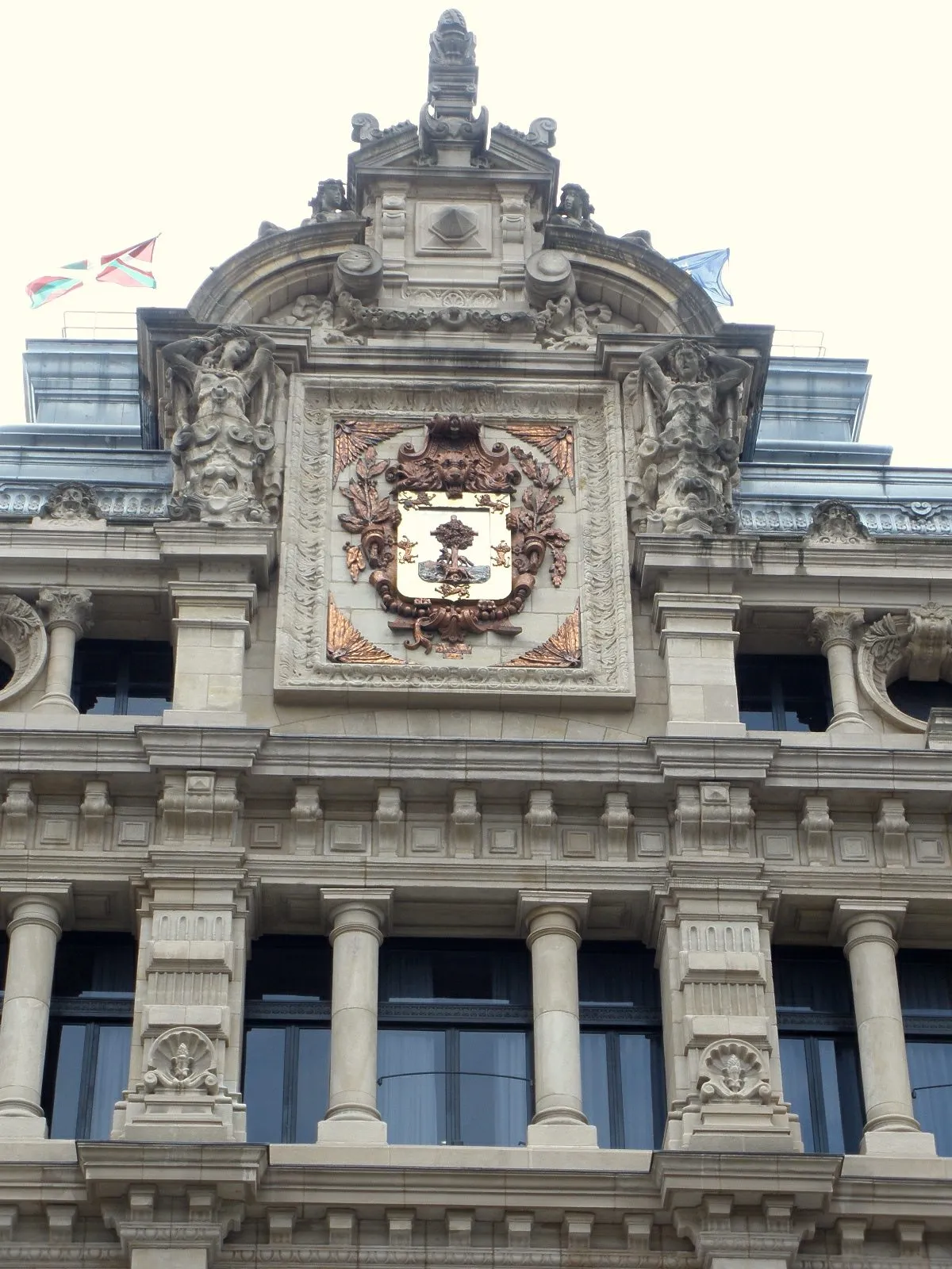 Ornate stone facade of the Bizkaia Provincial Council Palace showing a large decorative coat of arms flanked by sculptural figures, with columns, a pediment, and flags including the Basque flag at the top