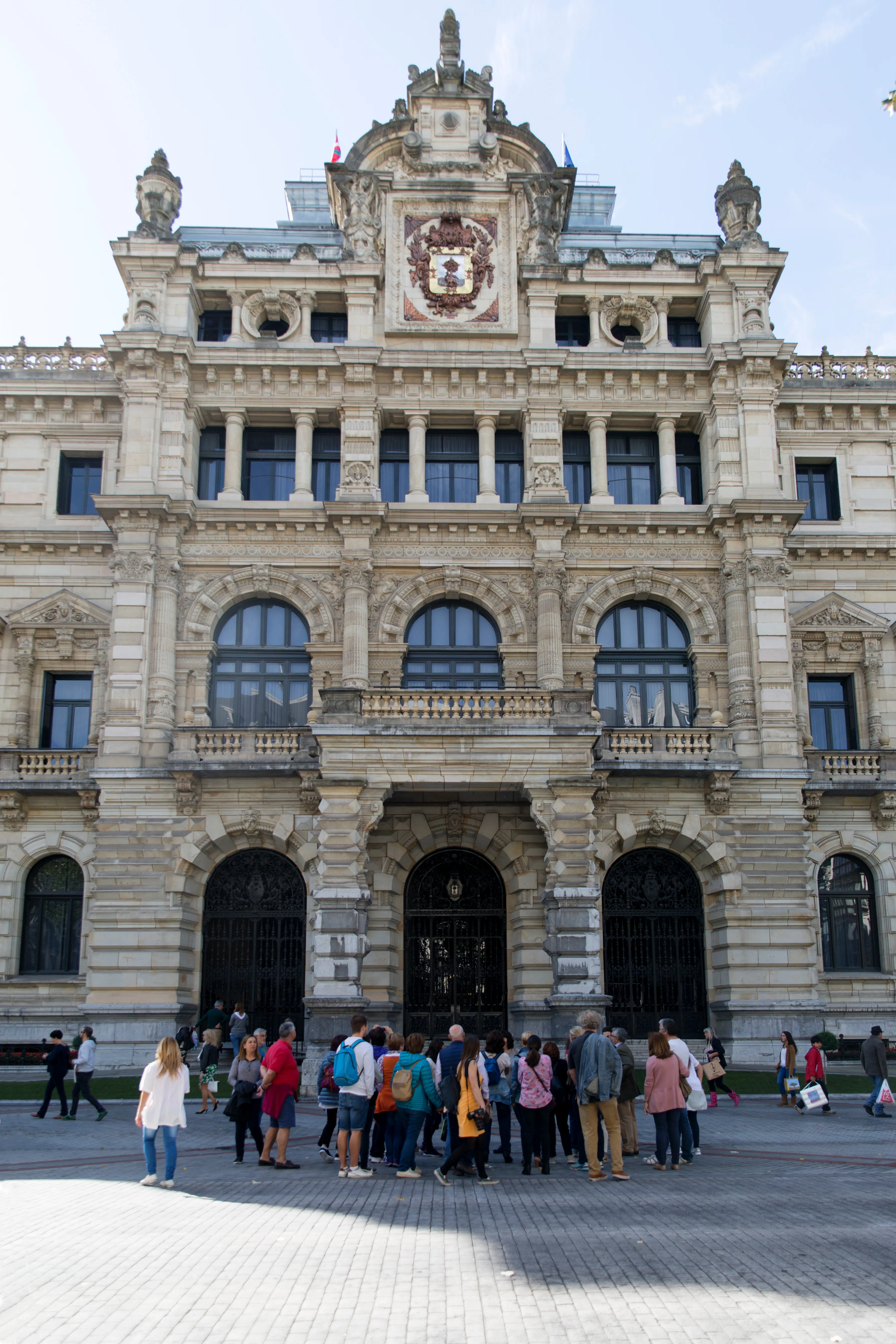 Ornate stone facade of the Bizkaia Provincial Council Palace with arched windows, decorative carvings, a central coat of arms, and visitors gathered in the plaza below