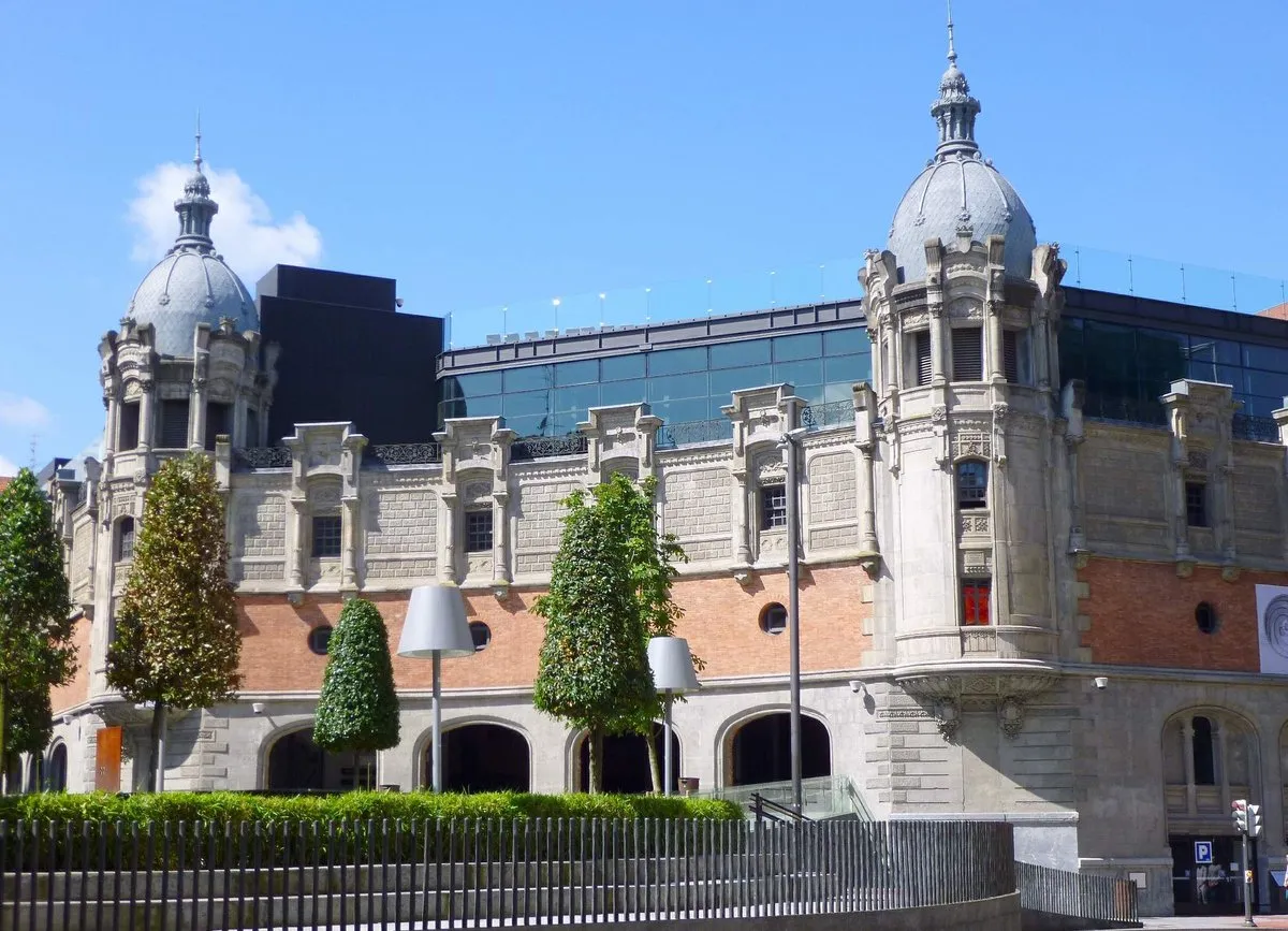 Exterior view of the Bizkaia Provincial Council Palace showing its ornate brick and stone facade, two prominent domed towers, and manicured trees in the foreground.