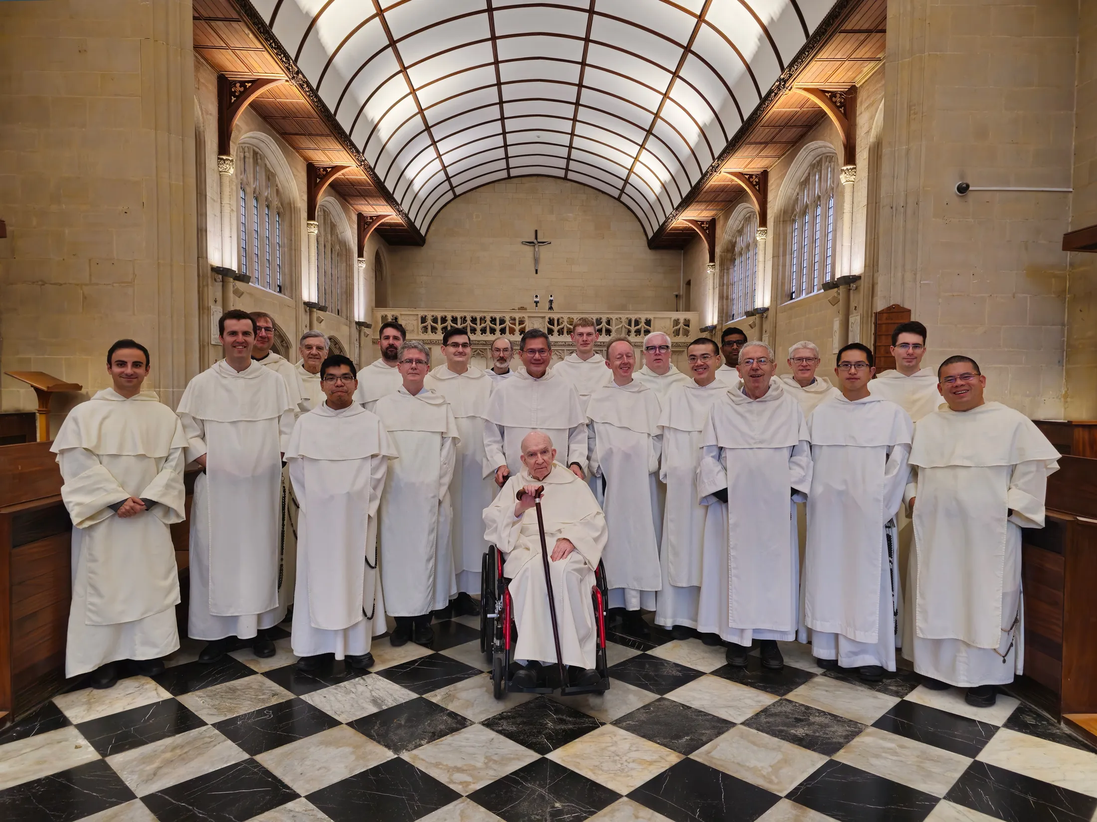 Grupa dominikanów w białych habitach pozująca wewnątrz kościoła Blackfriars w Oksfordzie, z czarno-białą szachownicą na podłodze, sklepieniem łukowym i ołtarzem widocznym w tle.