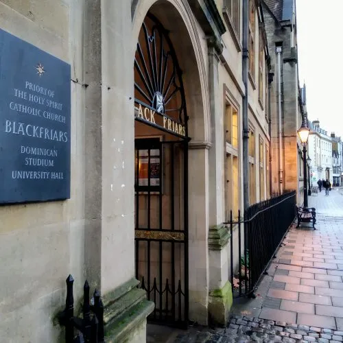Kamienna łukowa brama prowadząca do Blackfriars z niebieskim szyldem o treści 'Priory of the Holy Spirit Catholic Church Blackfriars Dominican Studium University Hall' oraz żelaznymi kratami